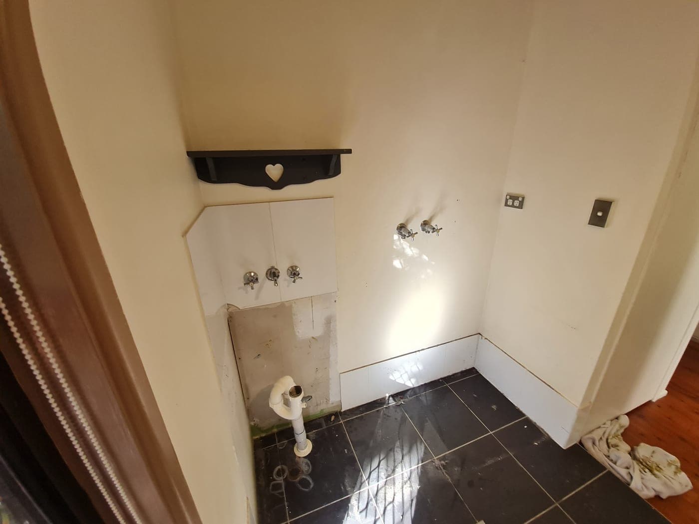 Old laundry stripped of appliances with bare wall taps and dated floor tiles