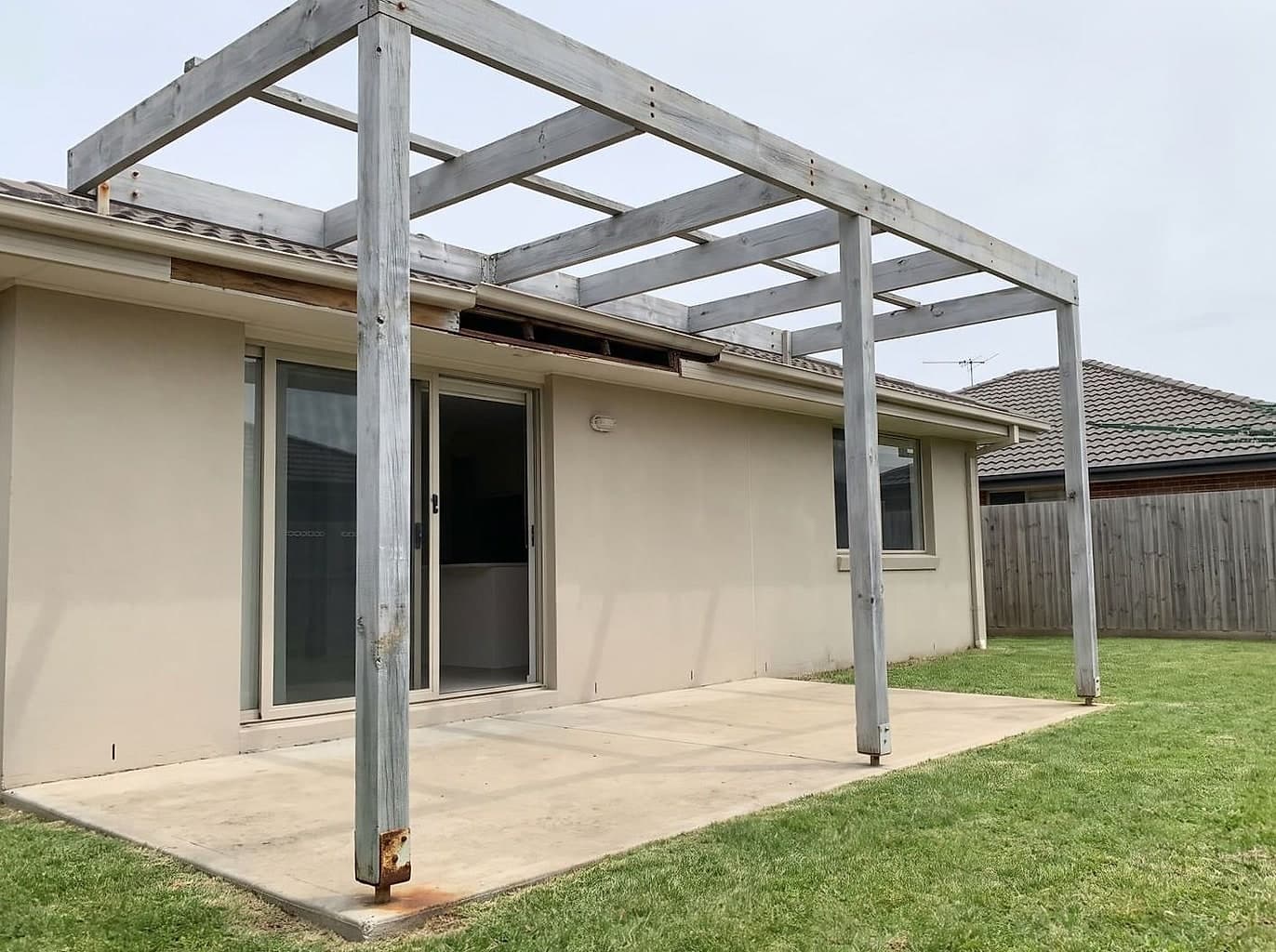 Weathered pergola over a concrete patio with its battens missing and a section of gutter taken down