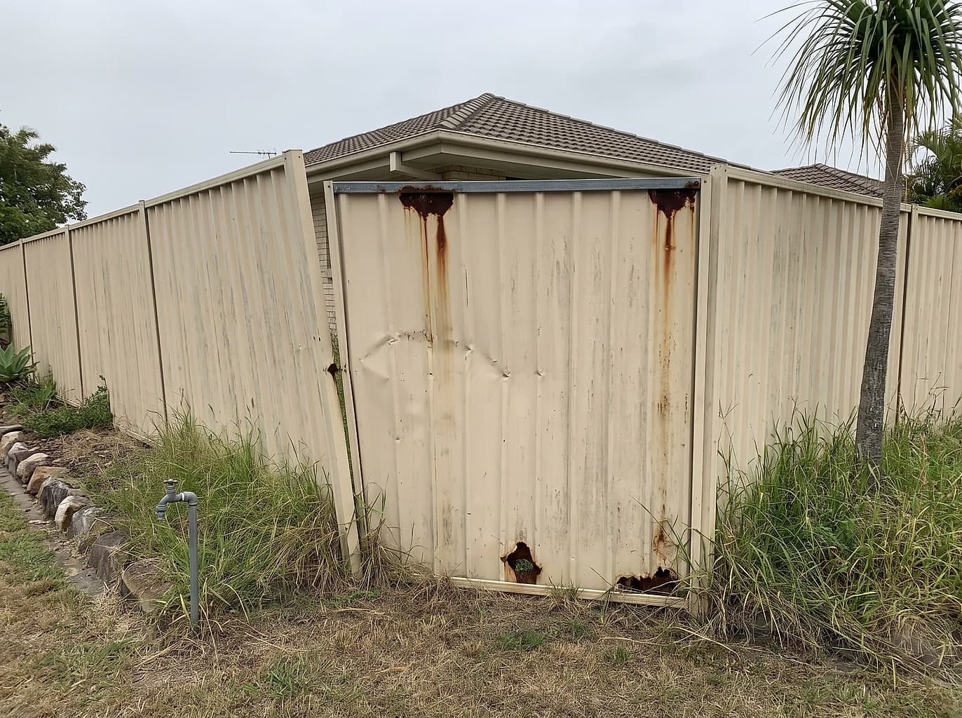 Faded colorbond fence dented, rusted through at the base and leaning out of line