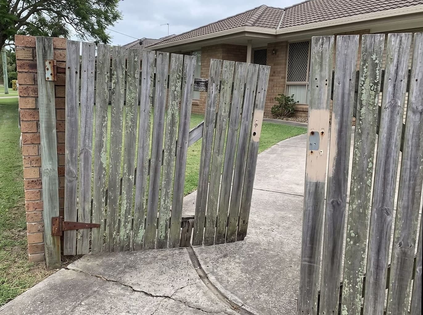 Timber front gate hanging from one hinge, palings missing and the latch taken off, dragging on the path