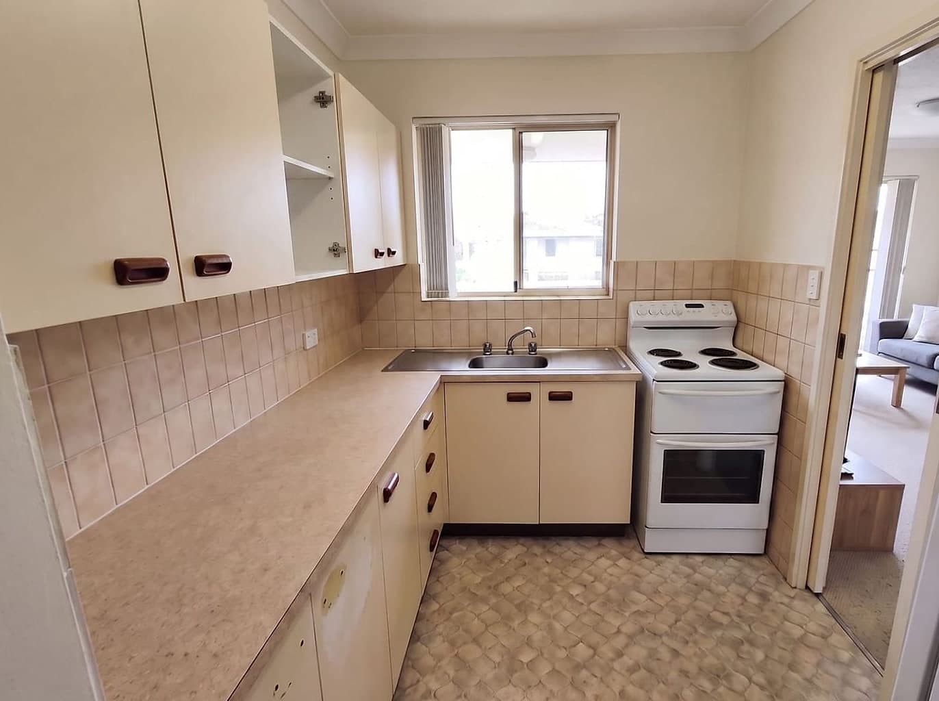 Dated unit kitchen with yellowed cream cupboards, worn laminate benchtop and an old freestanding stove
