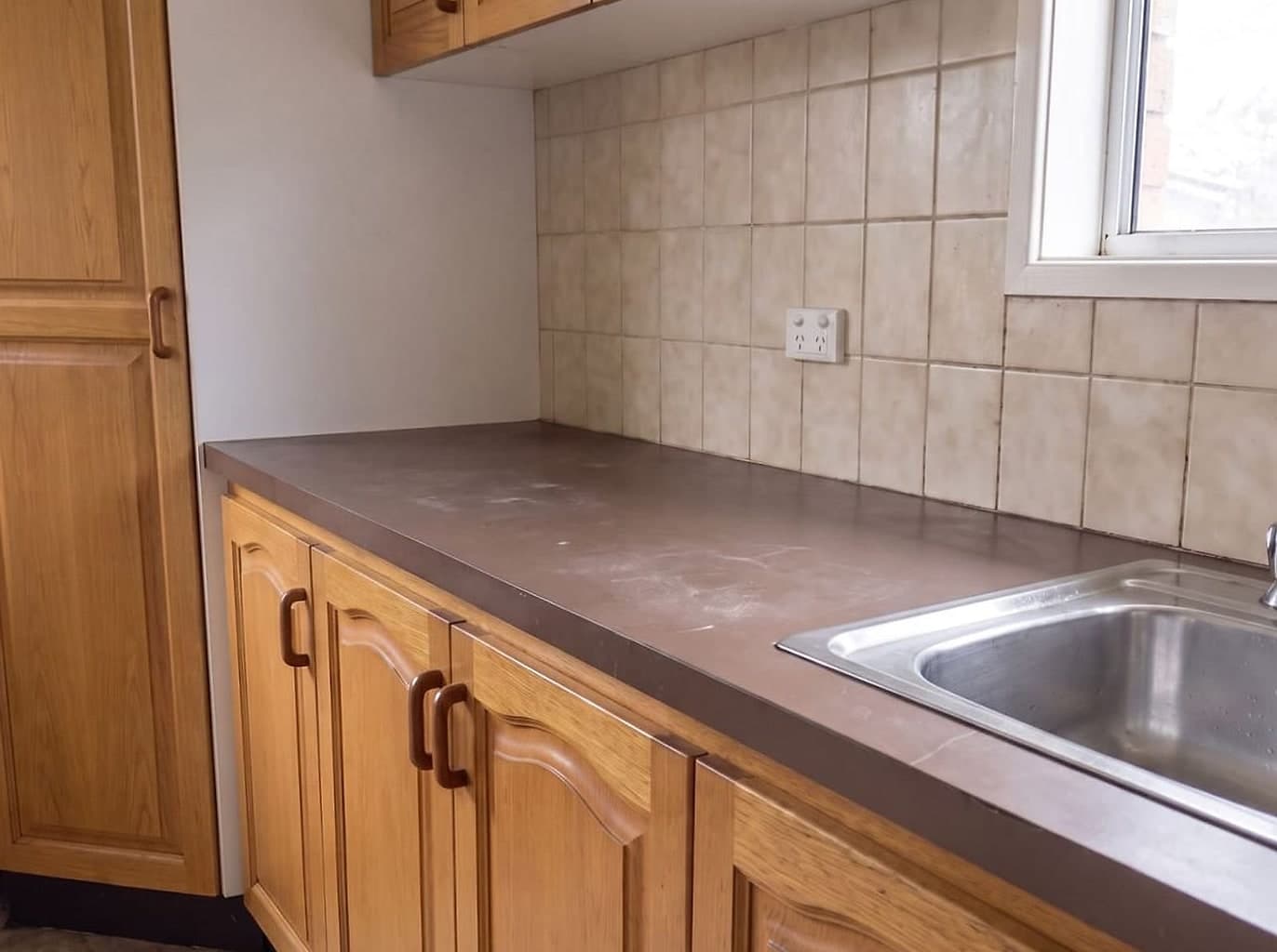 Scratched dark laminate benchtop with a swollen edge and a cracked beige tiled splashback