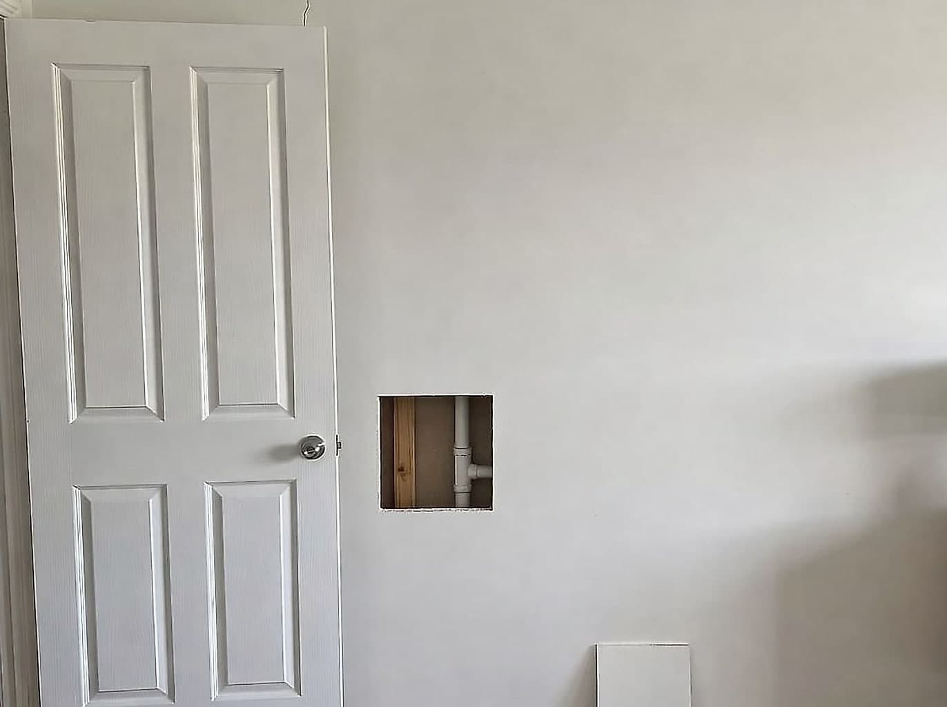 Wall with a square access cutout sawn through the plasterboard, a timber stud and a water pipe visible inside, and the offcut leaning below